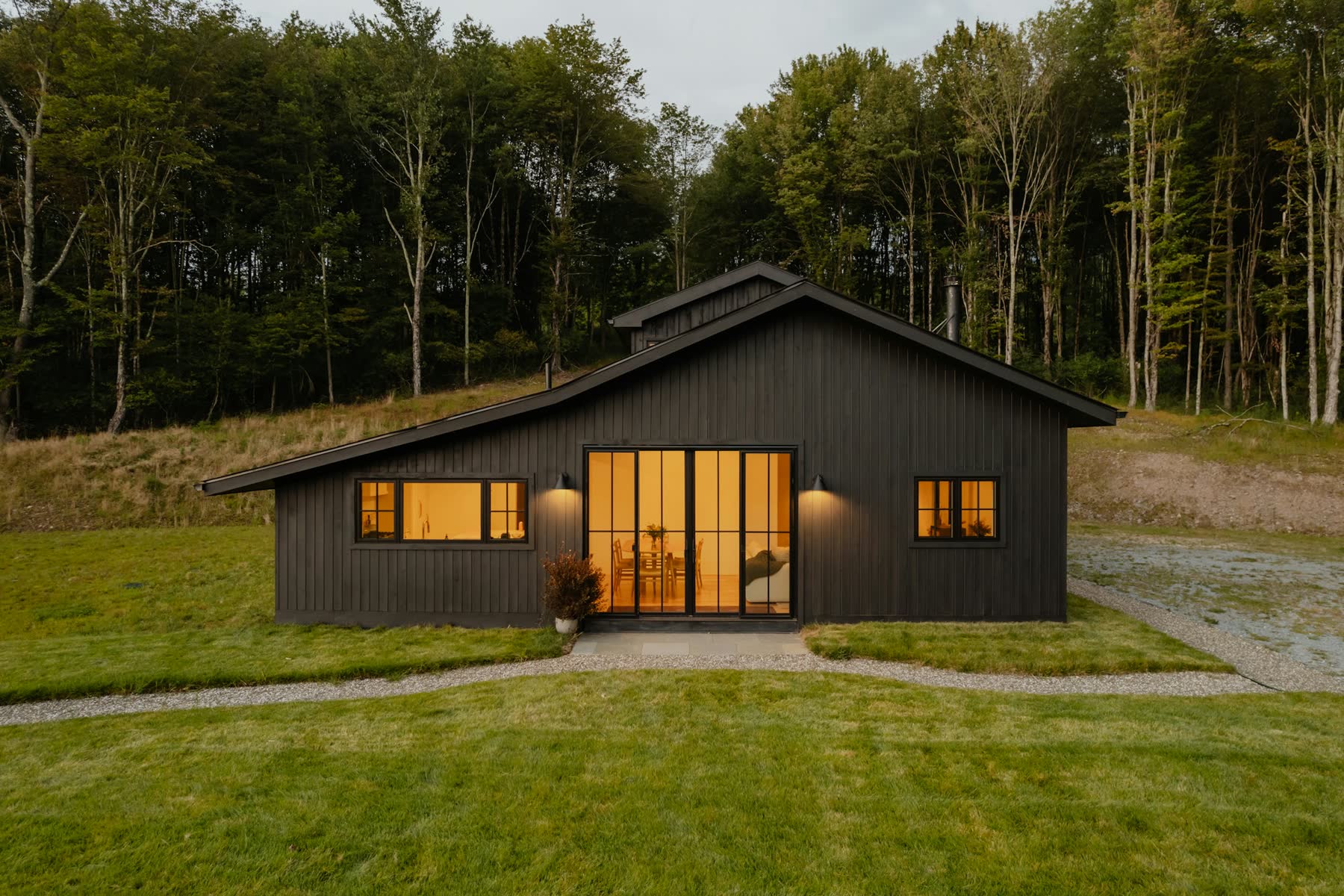 Casa de madera oscura con techo a dos aguas y ventanas iluminadas, sobre un claro de pasto